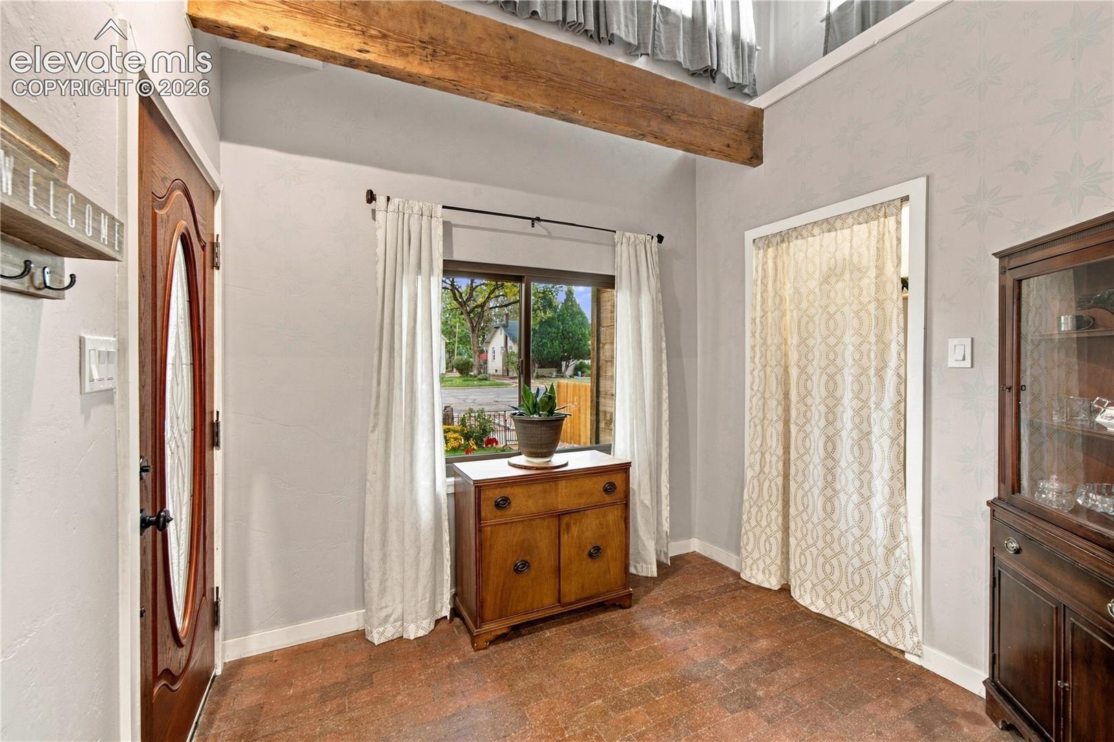 Entryway featuring a vaulted ceiling with exposed wood beam, patterned wall finish, textured tile flooring, and a solid wood door with an oval glass insert