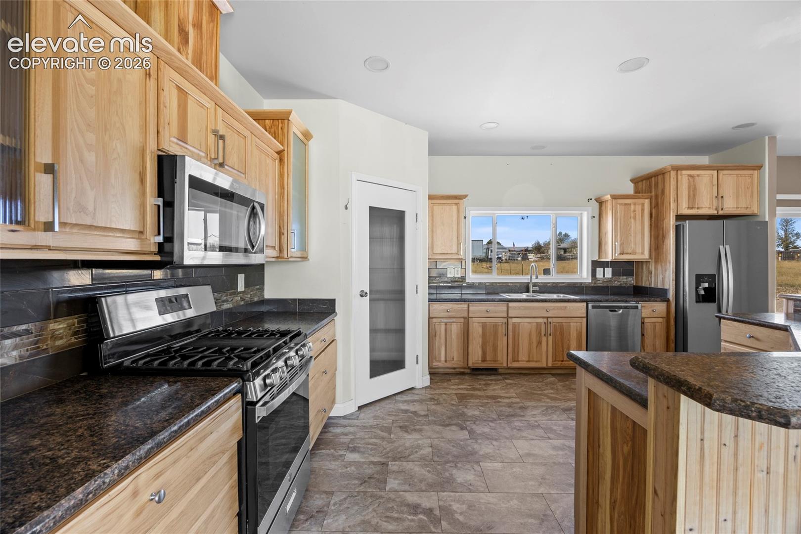 Kitchen featuring natural wood cabinetry, dark stone countertops, tile flooring, stainless steel appliances, and a pantry with a frosted glass door