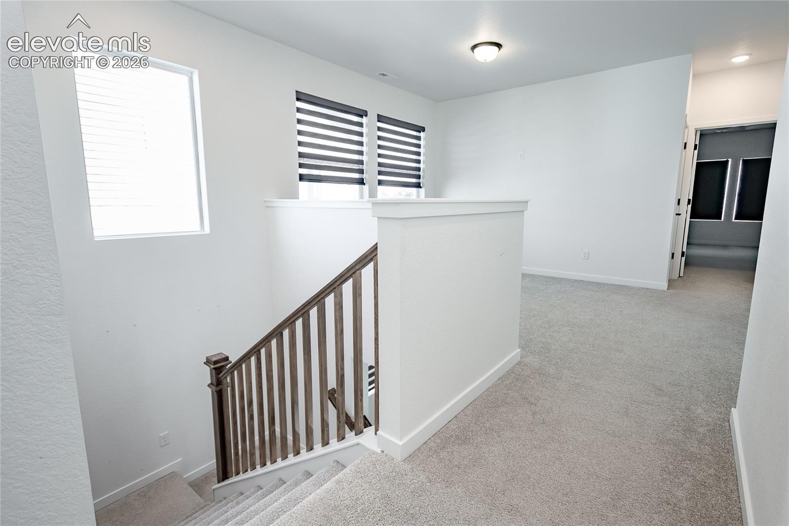 Open landing area featuring neutral carpeting, a staircase with a wood-finish handrail and vertical balusters, multiple windows with tiered window shades, and recessed ceiling lighting