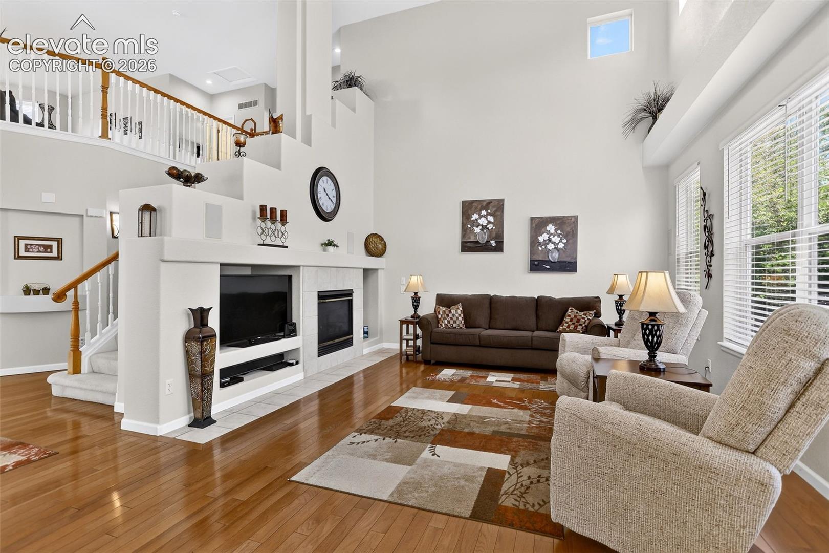 Expansive living area with high ceilings, large windows, wood-finish flooring, a built-in media center, and a fireplace
