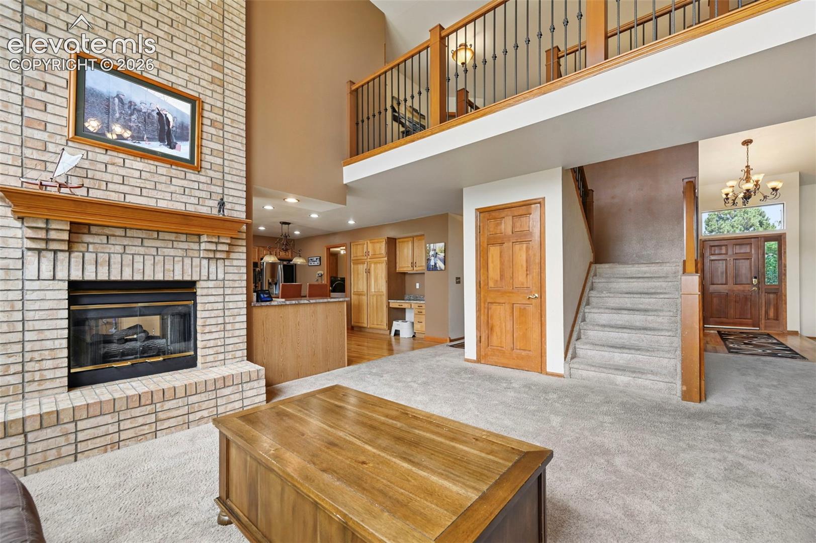 Brick fireplace with wood mantel, two-story ceiling, open mezzanine with wrought iron railing, wood entry door, and carpeted stairs