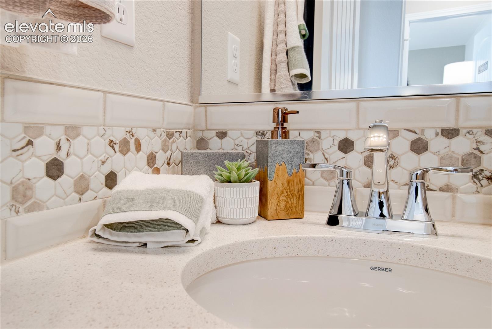 Bathroom vanity featuring a light-toned solid surface countertop, an undermount oval sink, chrome dual-handle faucet, and decorative hexagonal tile backsplash