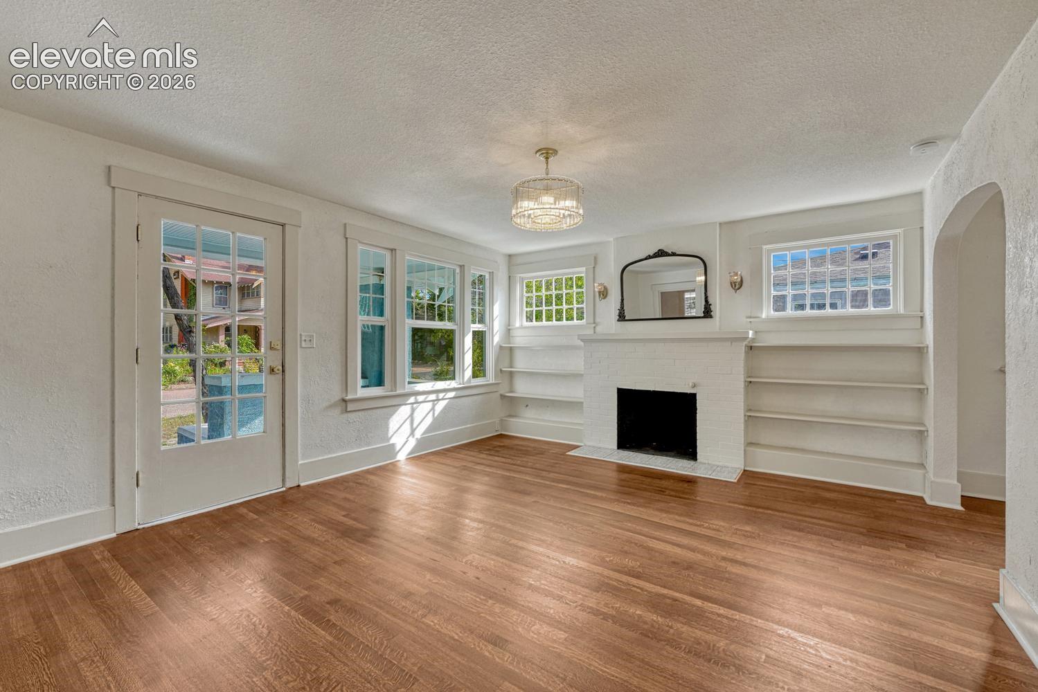 Hardwood flooring, white brick fireplace, built-in shelving, glass-paned door, and overhead chandelier