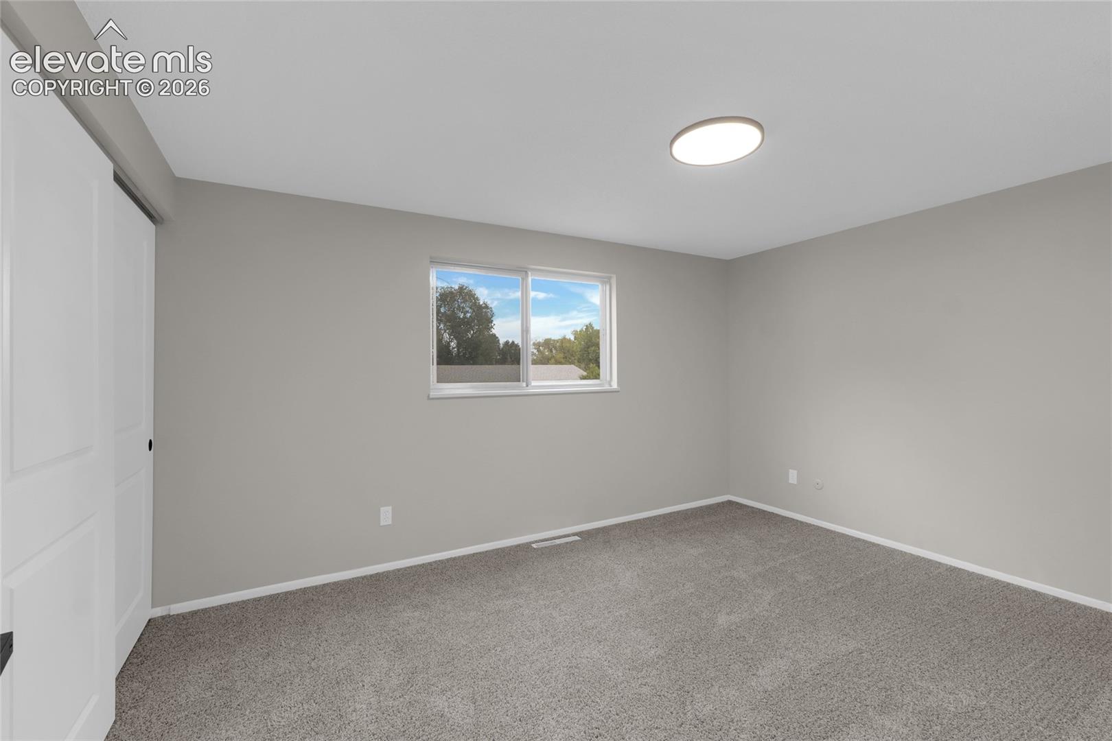 Neutral gray painted walls, window with white trim, gray carpet flooring, white sliding closet doors, and a flush-mount ceiling light