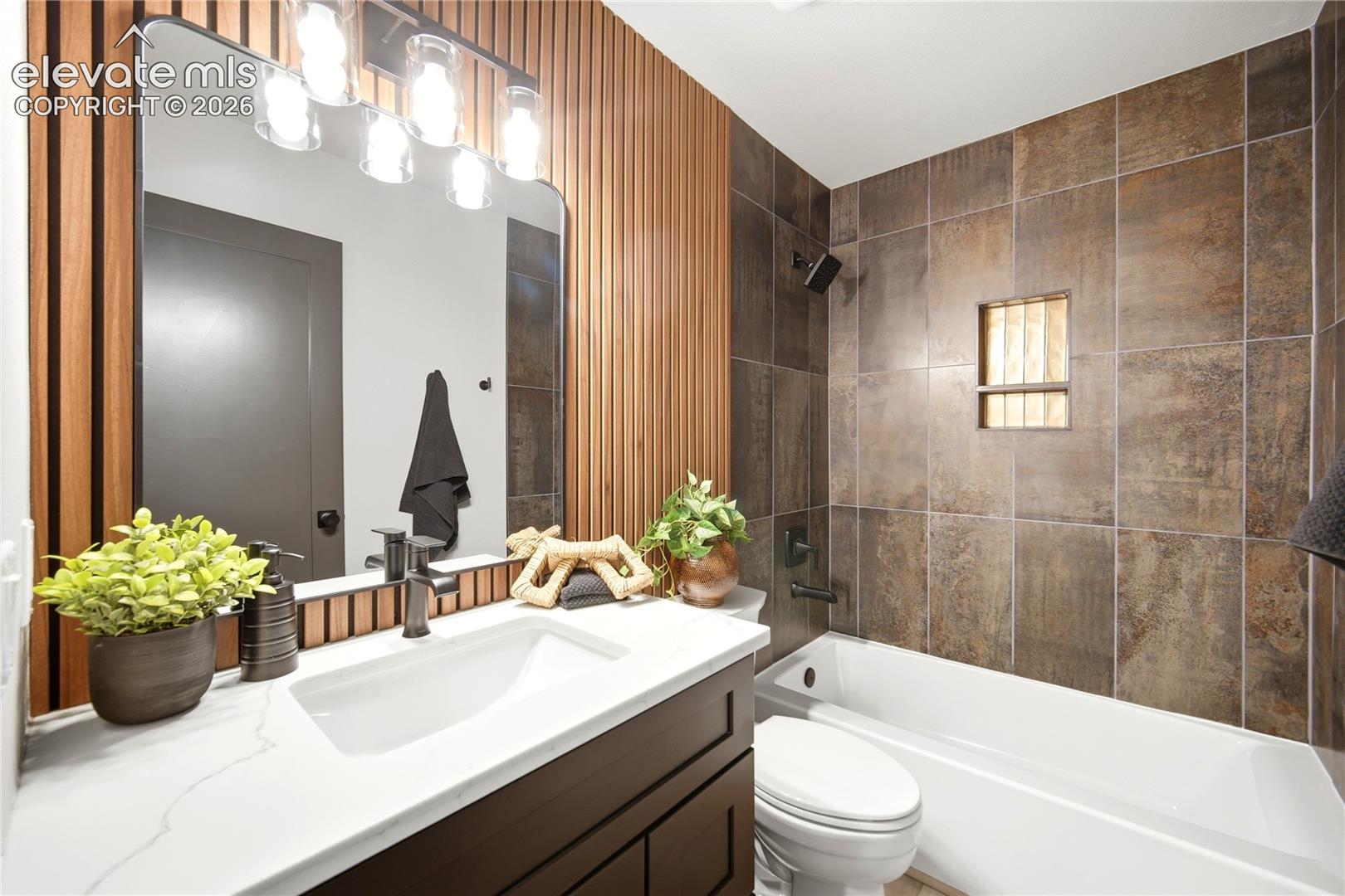Bathroom featuring a vanity with a white countertop and rectangular undermount sink, a vertical wood slat accent wall, a large vanity mirror with integrated multi-bulb lighting, and a bathtub surrounded by large-format textured wall tile with a built-in recessed shelf