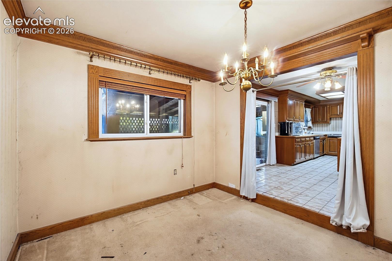 Wood-trimmed window, ornate crown molding, and a brass chandelier, with a spacious entry to the kitchen featuring wood cabinetry and tile flooring