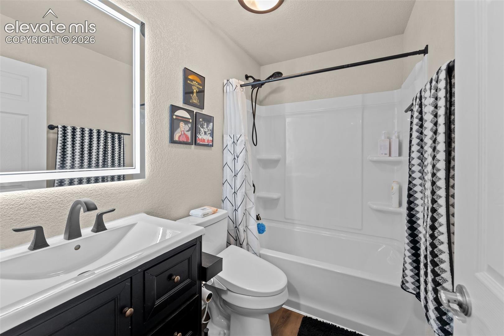 Backlit vanity mirror, black vanity with bronze hardware, matte black faucet, white toilet, and a white tub with shower
