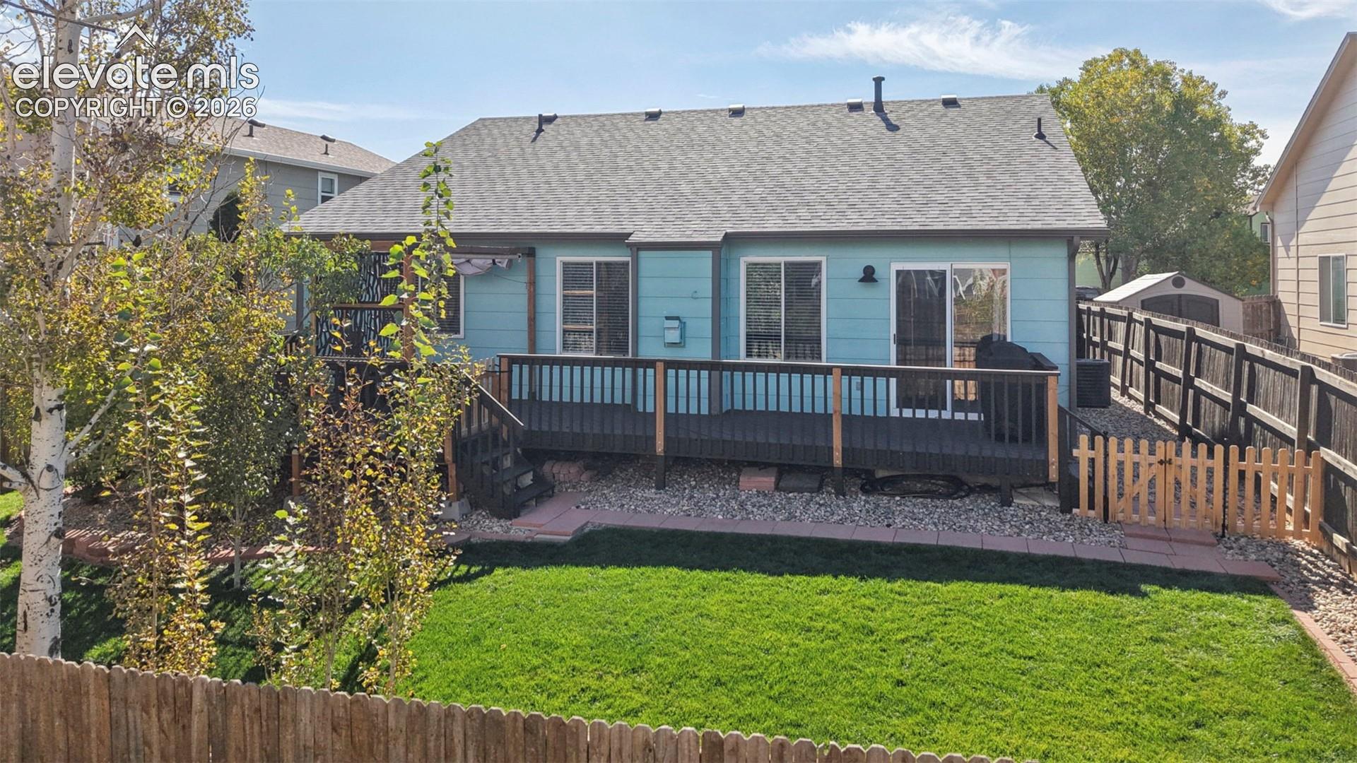 Exterior features include a horizontal lap siding facade, a raised wooden deck with railings, an enclosed backyard with a manicured lawn, stone landscaping borders, and a wood privacy fence