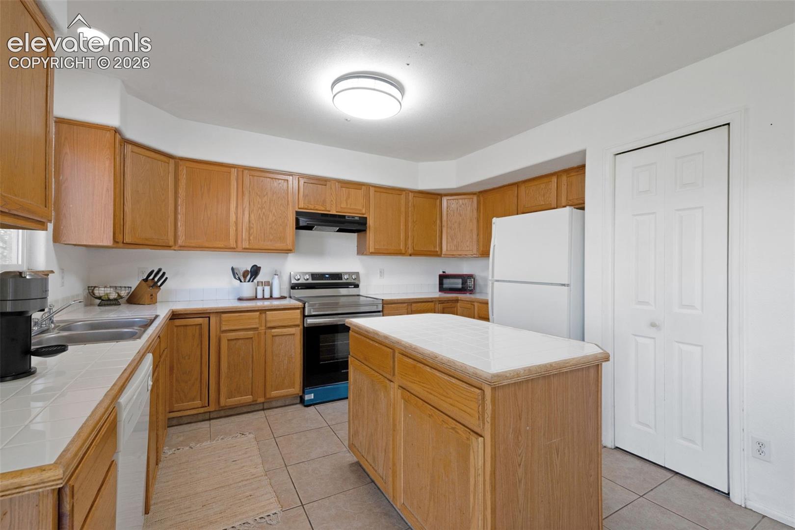 Kitchen featuring light wood cabinetry, a central island with a solid surface countertop, white tiled flooring, and a comprehensive appliance suite including a stainless steel range with overhead ventilation