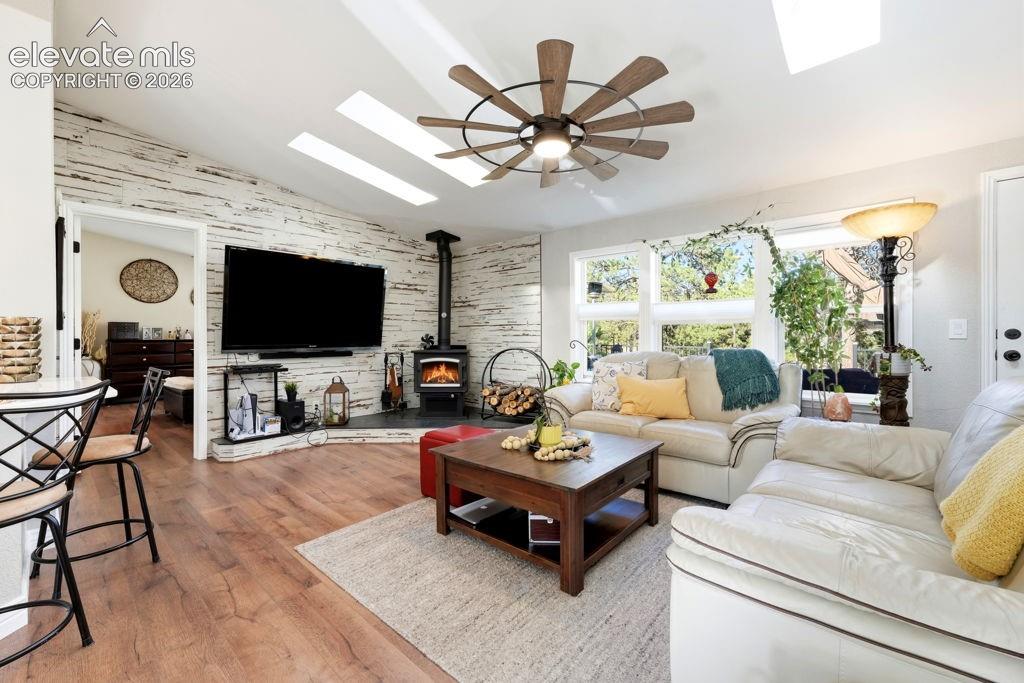 Vaulted ceiling with skylights, wood-finish flooring, accent wall, wood-burning stove, and ceiling fan