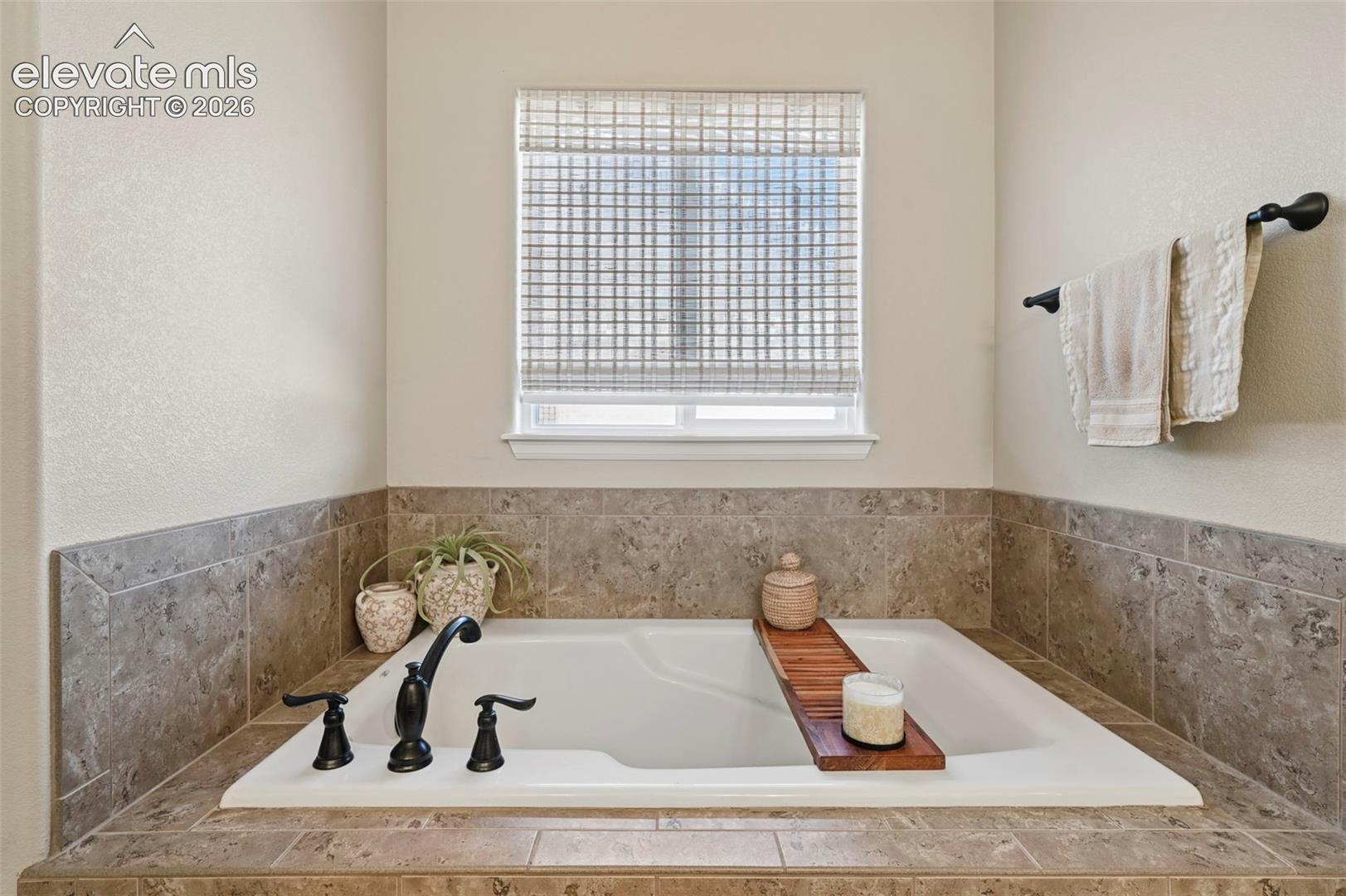 Built-in soaking tub with a tiled surround, black hardware, and an adjacent window with woven blinds