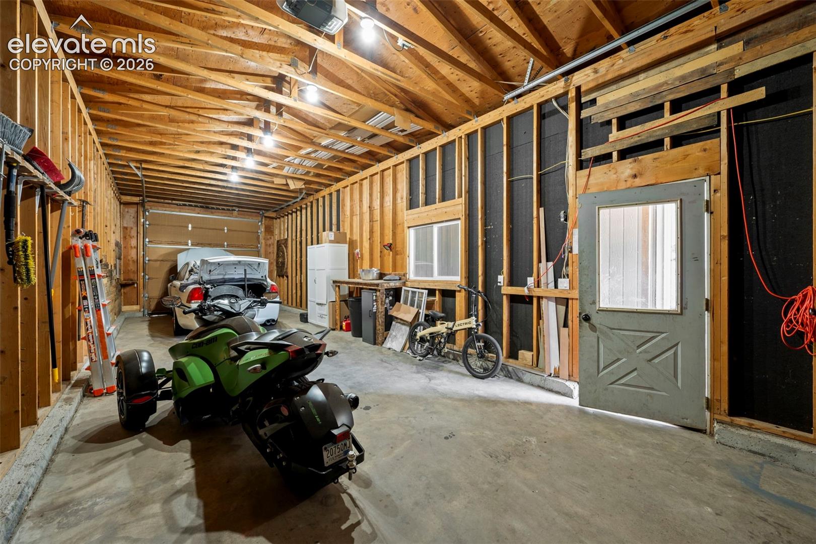 Unfinished garage featuring exposed wood framing, concrete flooring, overhead lighting, and a solid-panel door with a glass inset