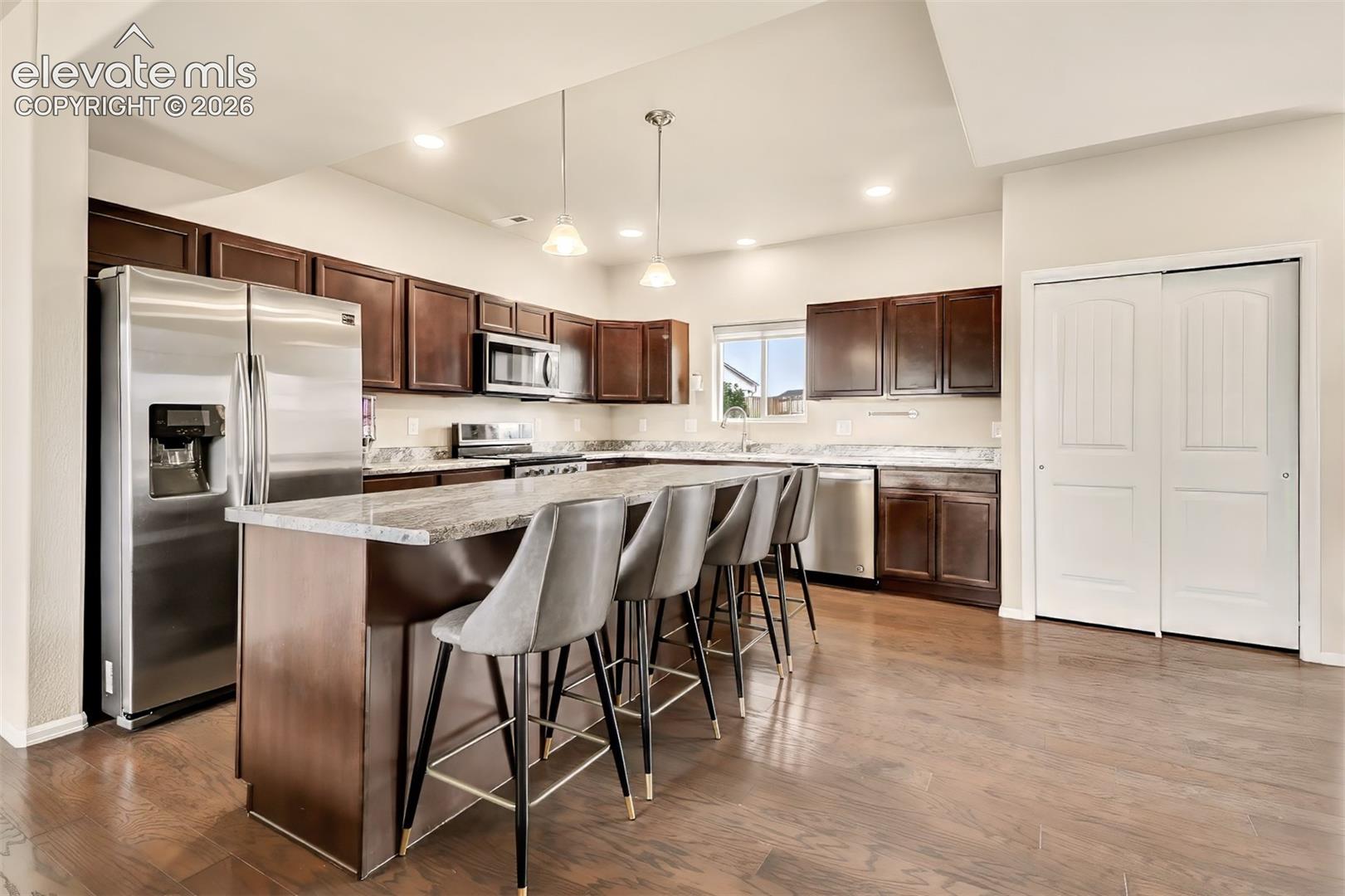 Kitchen featuring dark wood cabinetry, stainless steel appliances, stone-finish countertops, wood-finish flooring, and a center island with dual pendant lighting