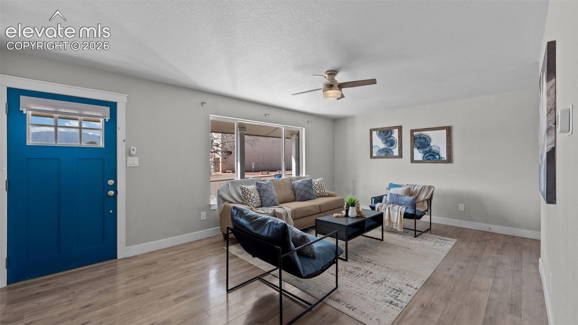 Wood-finish flooring throughout, bright blue entry door with transom window, ceiling fan with integrated lighting, and multiple large windows