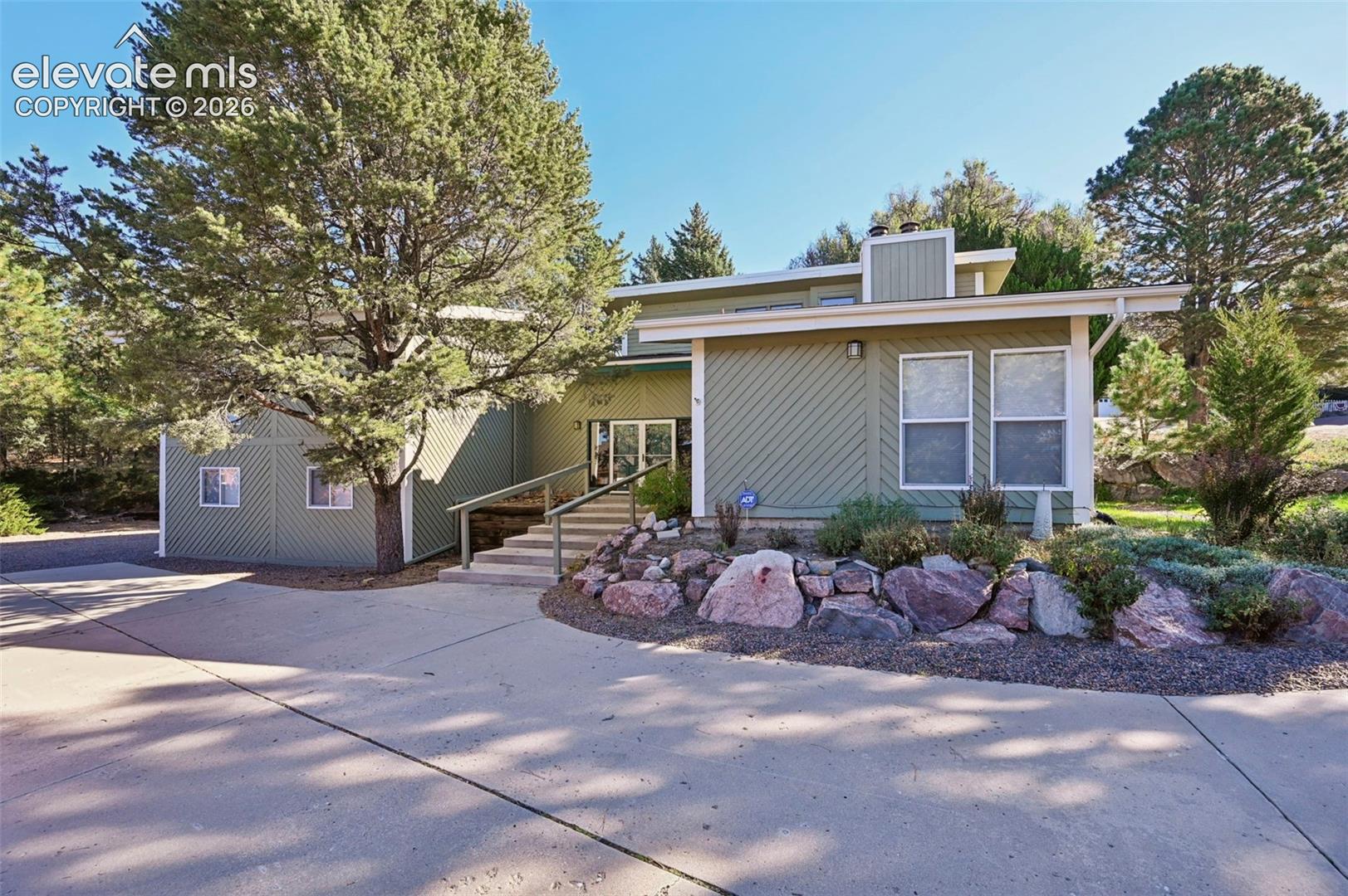Split-level exterior featuring diagonal wood siding, concrete driveway, stone rockery landscaping, and a railed staircase leading to the entryway