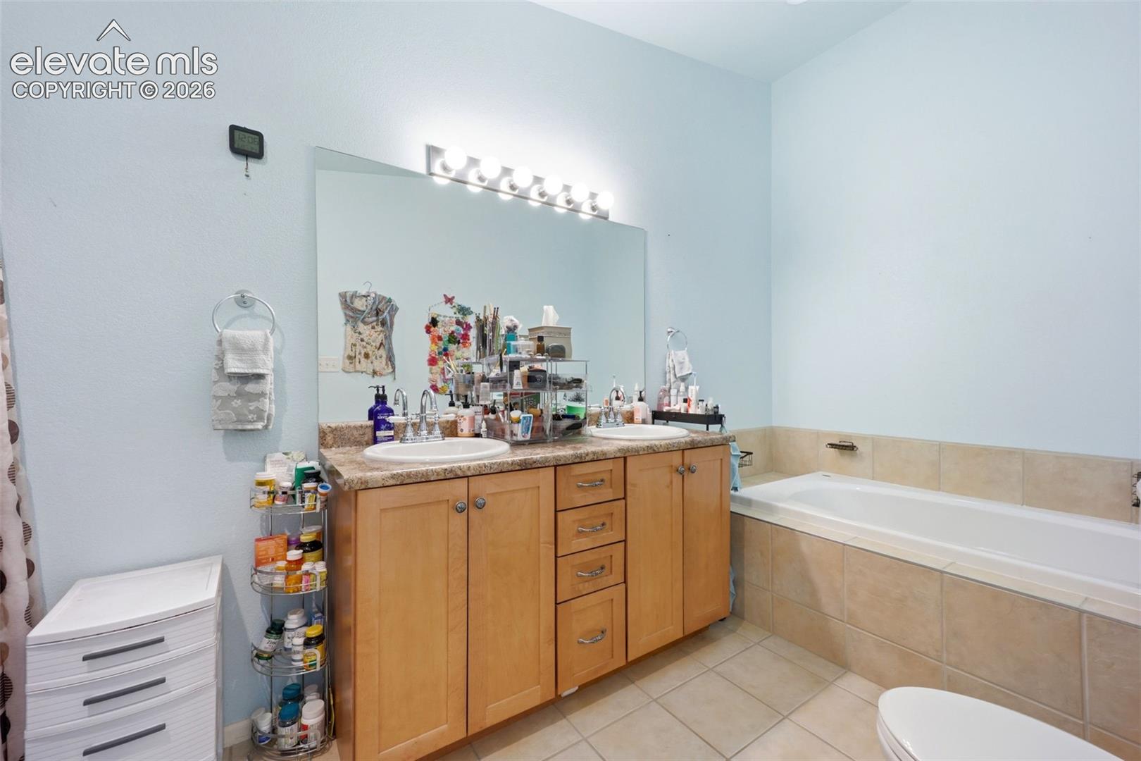 Bathroom featuring a dual vanity with light wood cabinetry, a built-in bathtub with tiled surround, a large wall mirror, and tiled flooring