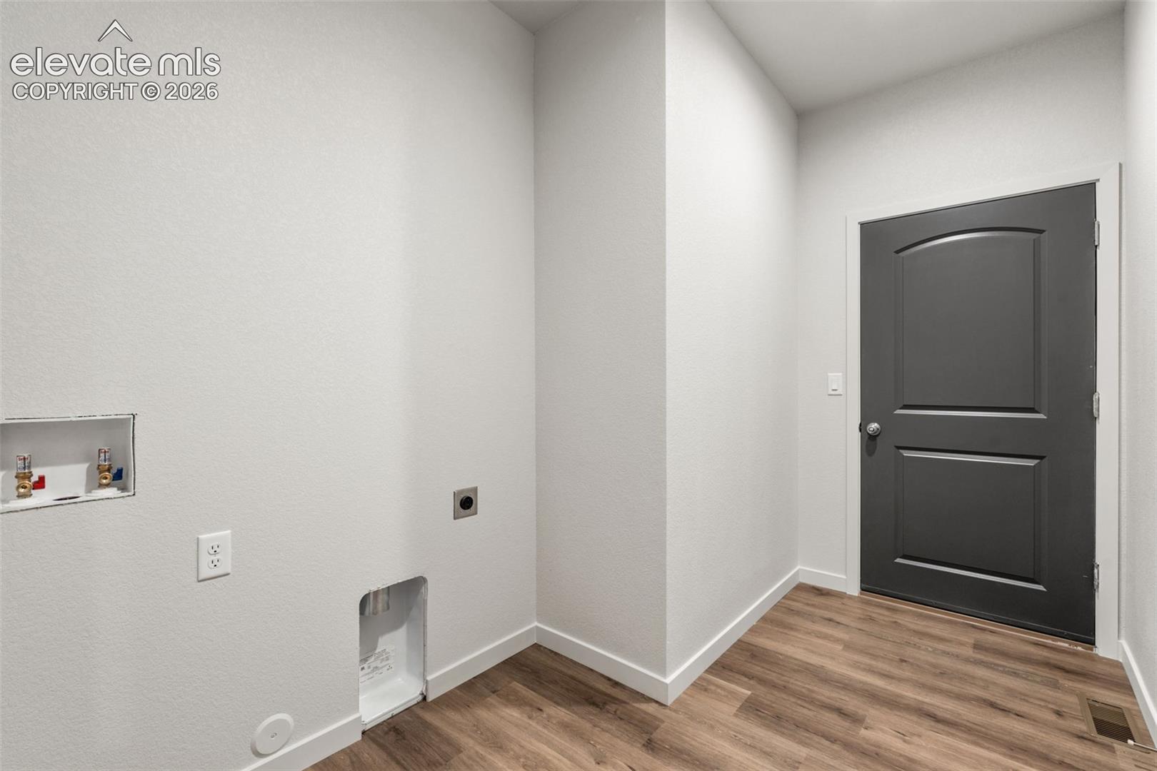 Utility area featuring recessed wall-mounted plumbing hookups, wood-finish flooring, and a dark-toned interior door with an arched-panel design