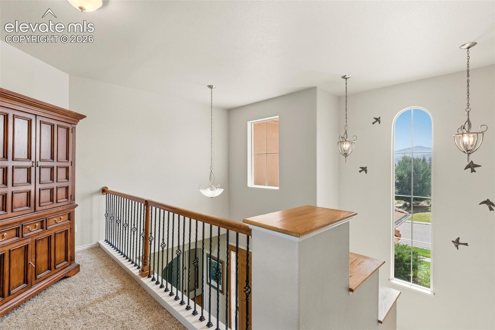 Upper-level landing featuring a wrought iron and wood-finish railing, arched windows, recessed lighting, and pendant light fixtures