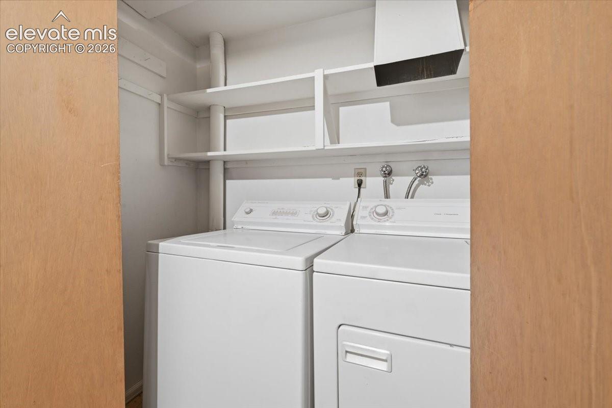 Laundry closet featuring white top-load washer and dryer, dual overhead white shelving, and dual wood-finish bi-fold doors