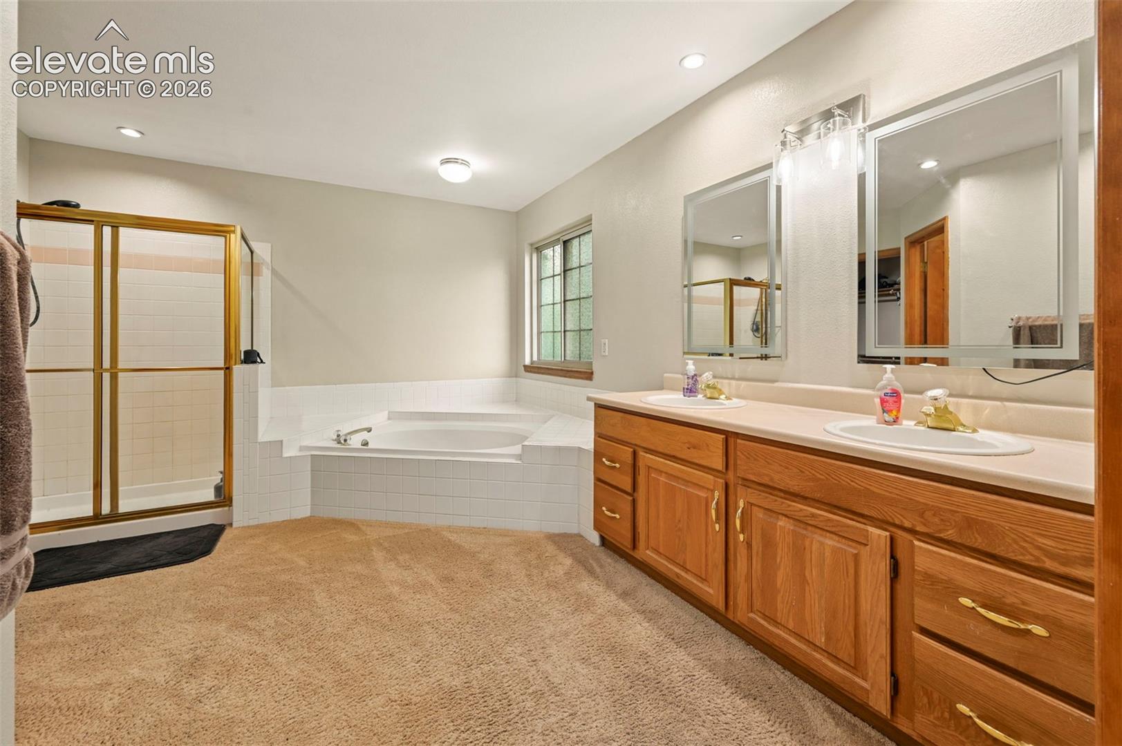 Dual vanity with wood-finish cabinetry, built-in soaking tub, separate shower enclosure with gold-tone framing, and recessed lighting