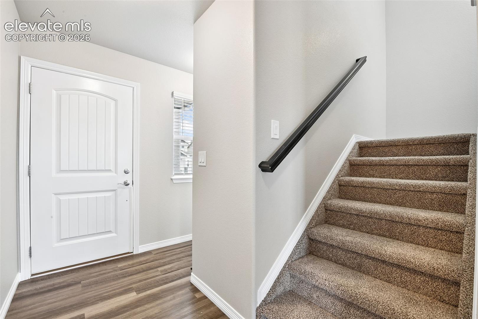 Entryway featuring a white paneled door, wood-finish flooring, a single-hung window, and carpeted stairs with a mounted metal handrail