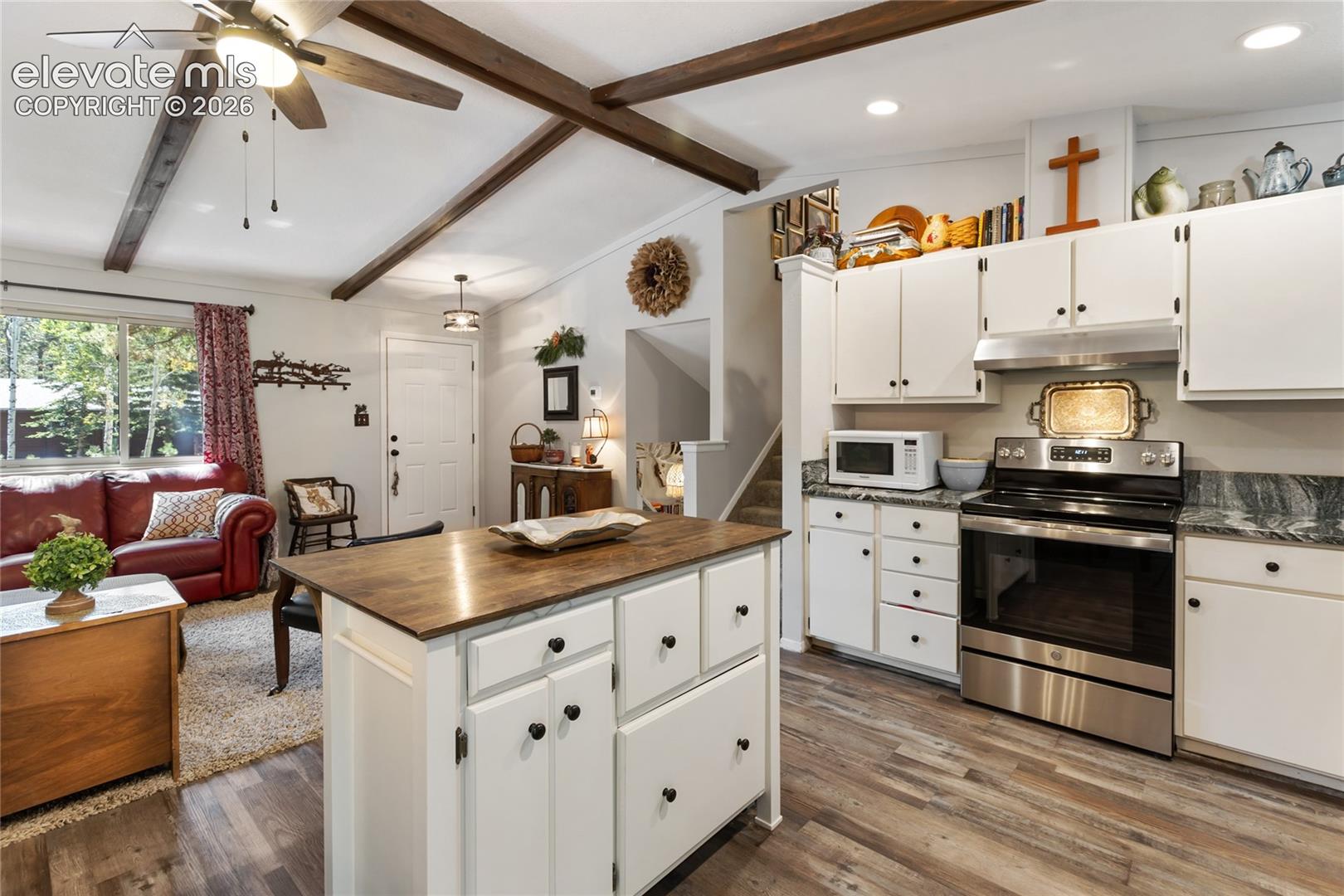 Vaulted ceiling with exposed wood beams, wood-finish flooring, white cabinetry with black hardware, stainless steel range and microwave, and a kitchen island with wood-finish countertop