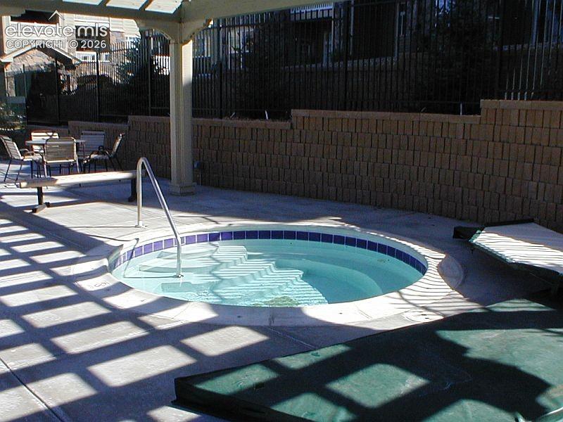 In-ground circular spa with tile border and integrated steps, concrete patio surface, overhead wooden pergola, and segmented stone retaining wall
