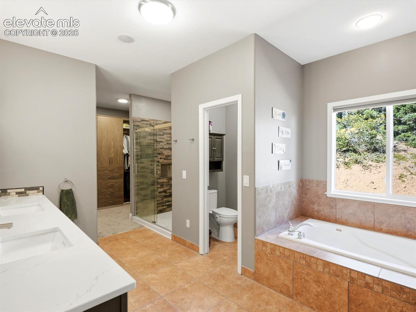 Double vanity with a white countertop and rectangular undermount sinks, stone-tile flooring, soaking tub with a tiled surround, glass-enclosed shower with stone-tile walls, and built-in wood-finish cabinetry