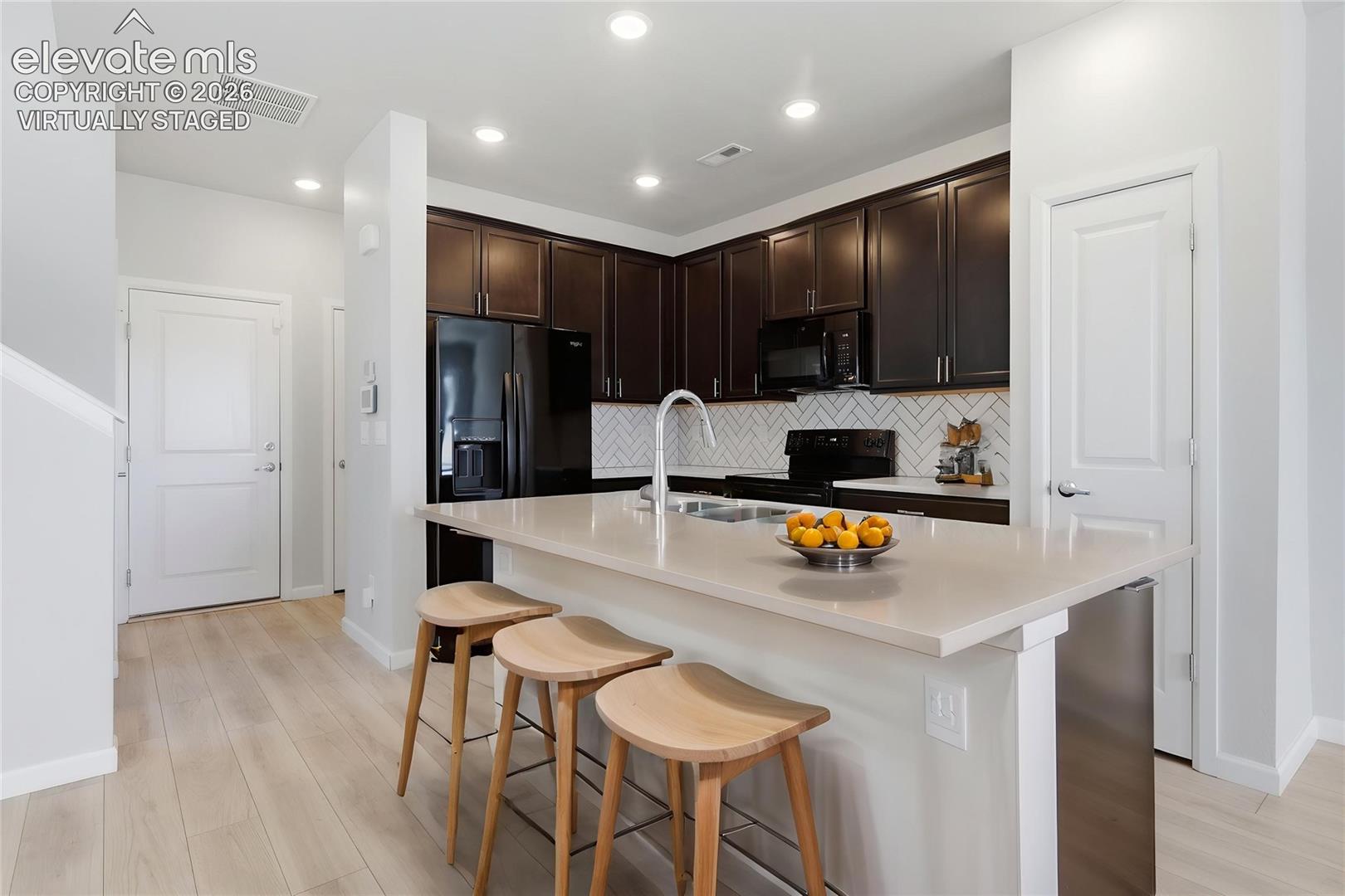 Kitchen featuring dark wood cabinetry, a light-toned countertop island with an undermount sink, black appliances, and a white subway tile backsplash in a herringbone pattern -- virtually staged.