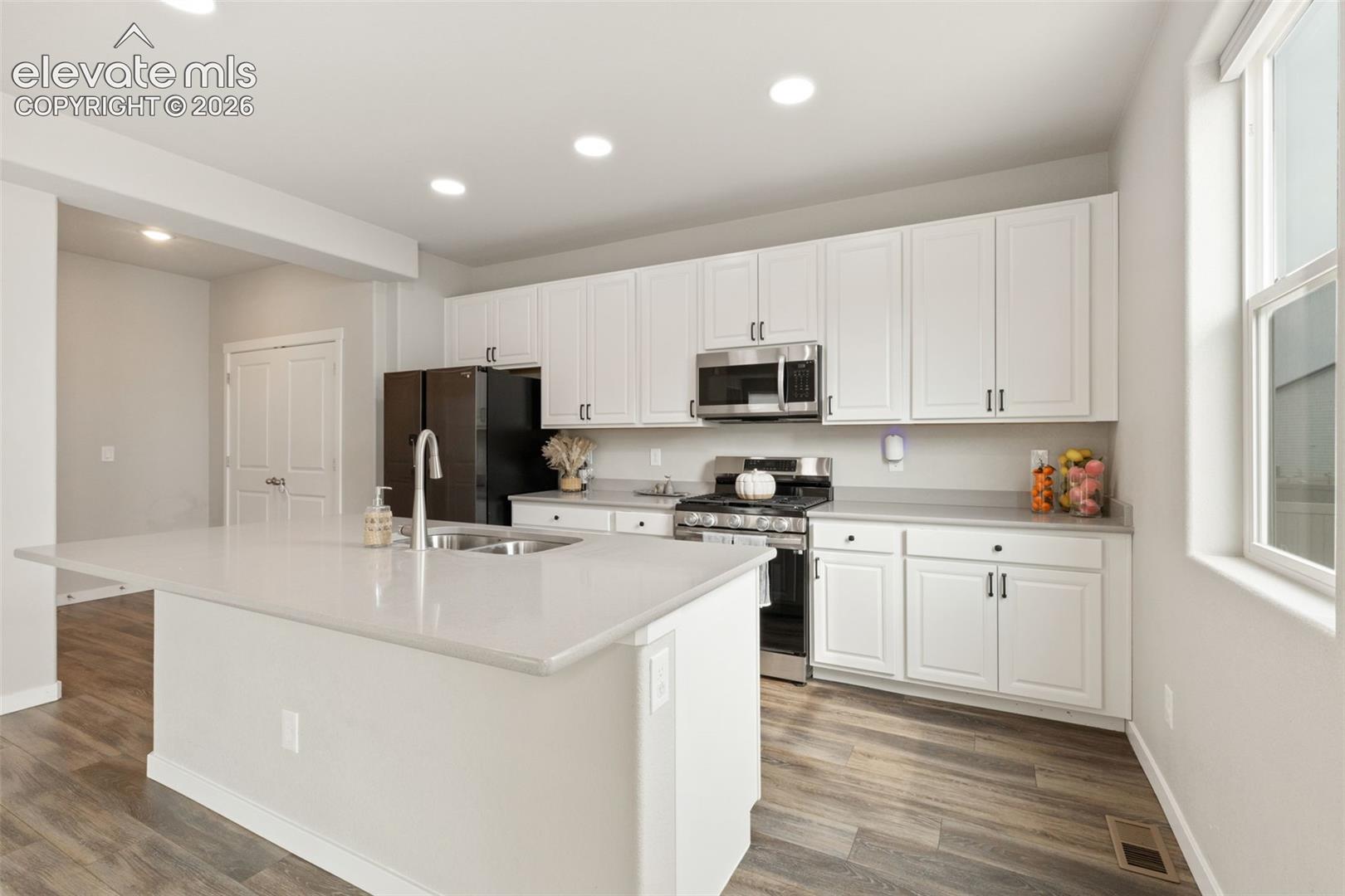 Modern kitchen featuring a large island with an undermount sink, light-toned countertops, white shaker cabinetry, stainless steel appliances, gas stove, and wood-finish flooring
