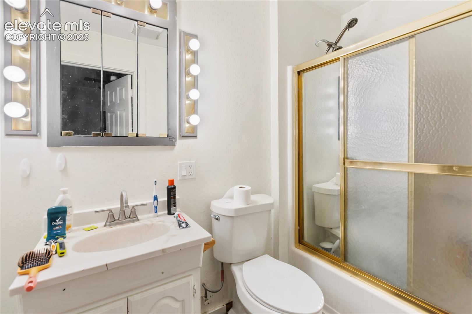 Bathroom featuring a white vanity cabinet with a stone countertop, integrated oval sink, and a wall-mounted mirrored medicine cabinet flanked by multi-bulb light fixtures, alongside a bathtub with a sliding glass door and gold-finish frame