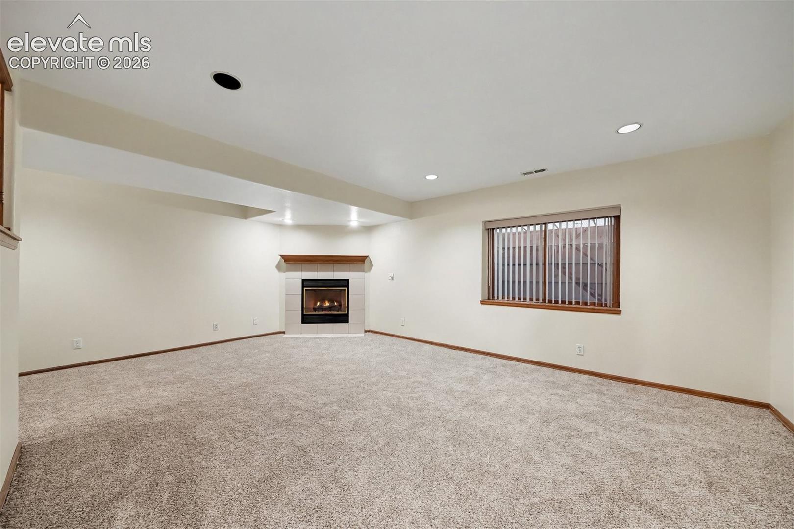 Expansive carpeted room featuring a built-in fireplace with a tile surround and wood mantel, recessed lighting, and a window with blinds