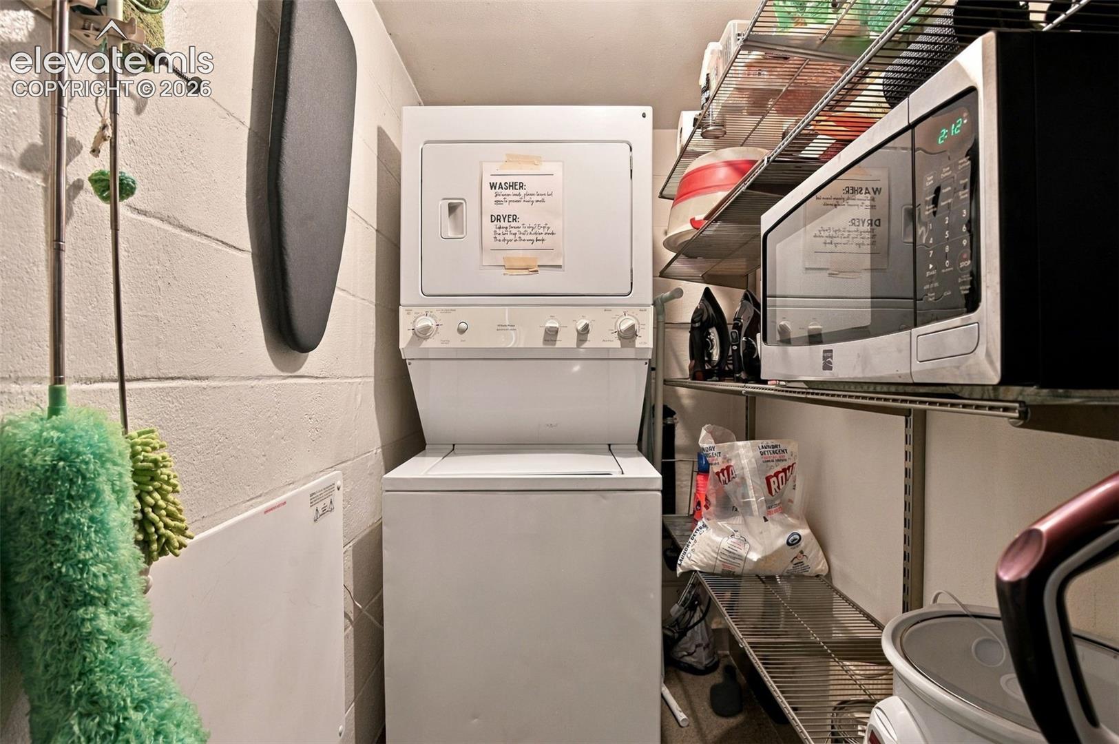 Stacked laundry unit featuring white appliance finish, complemented by integrated metal wire shelving and masonry wall construction
