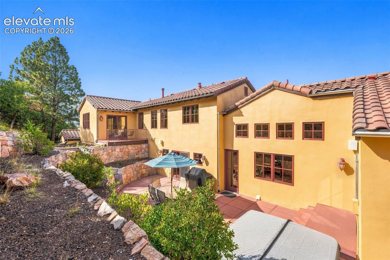 Multi-level exterior featuring stucco siding, tile roof, multiple windows, an upper-level balcony, and a lower-level patio with stone retaining walls
