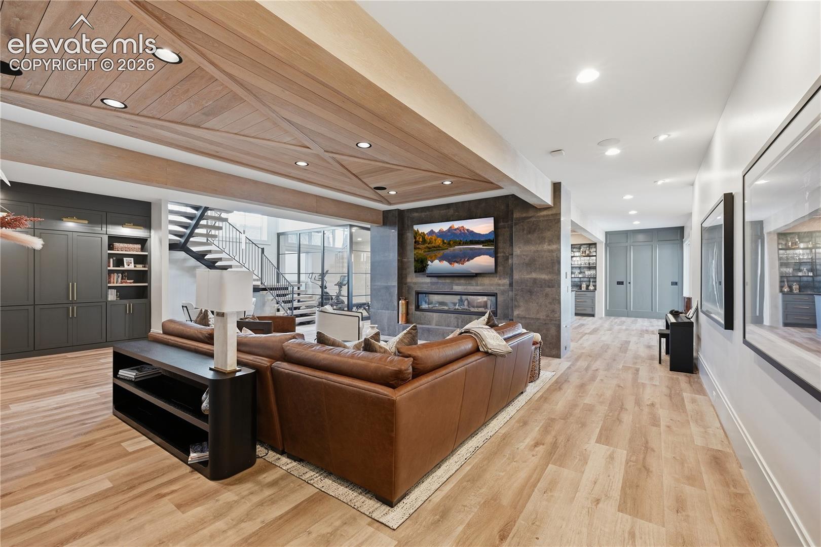 Living space featuring a wood-plank ceiling with decorative beam inlay, a floor-to-ceiling stone-clad fireplace with an integrated linear firebox, wood-finish flooring, custom charcoal-toned built-in cabinetry, and a floating metal staircase with glass railing