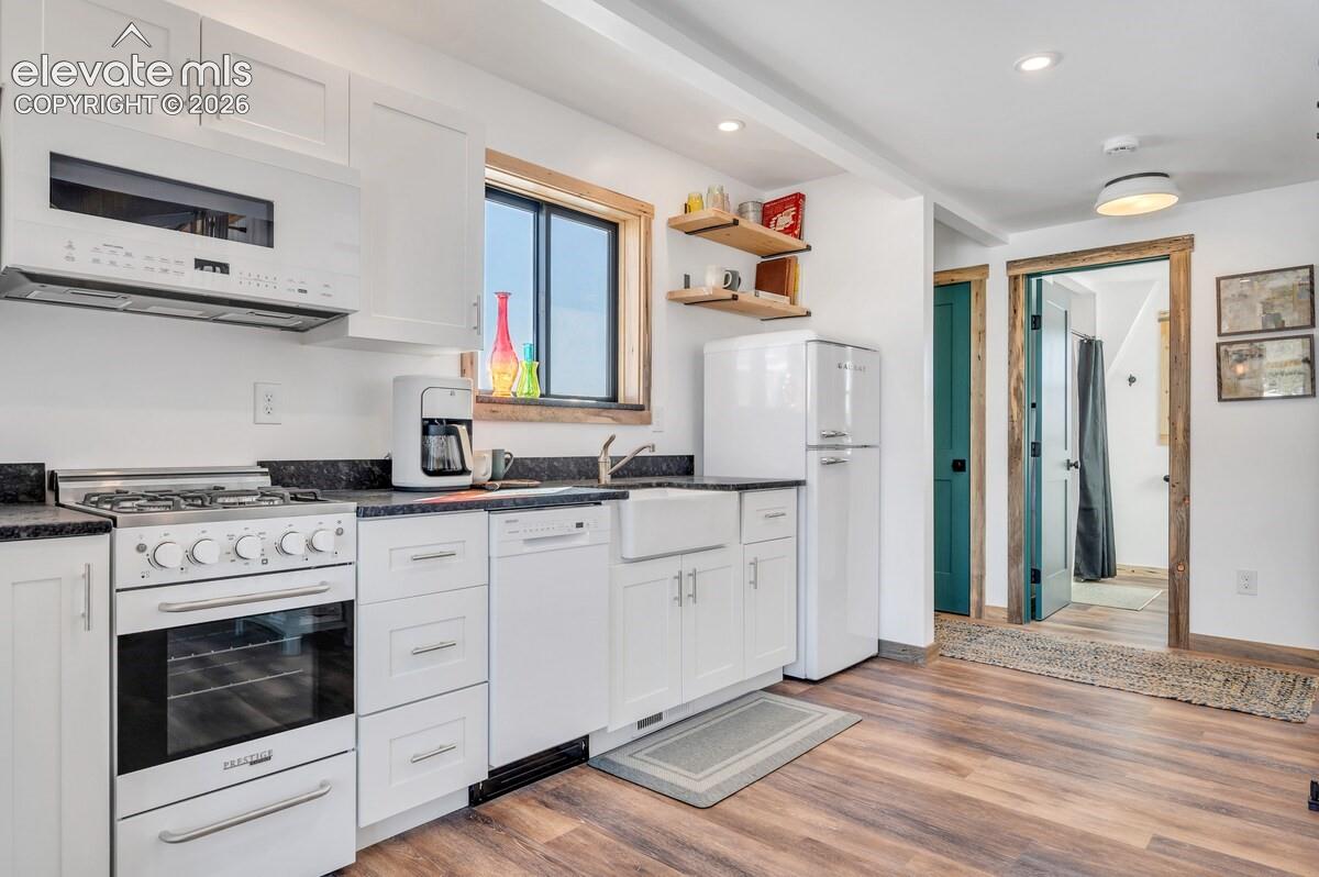 Kitchen featuring white shaker-style cabinetry, dark stone-finish countertops, an apron-front farmhouse sink, gas range, and wood-finish flooring
