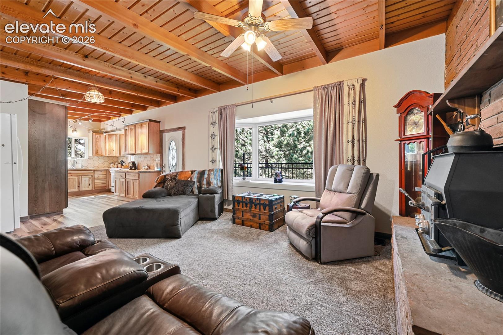 Exposed wood beam ceiling, bay window, wood-burning stove with brick surround, wood-finish flooring in kitchen, and light-toned cabinetry