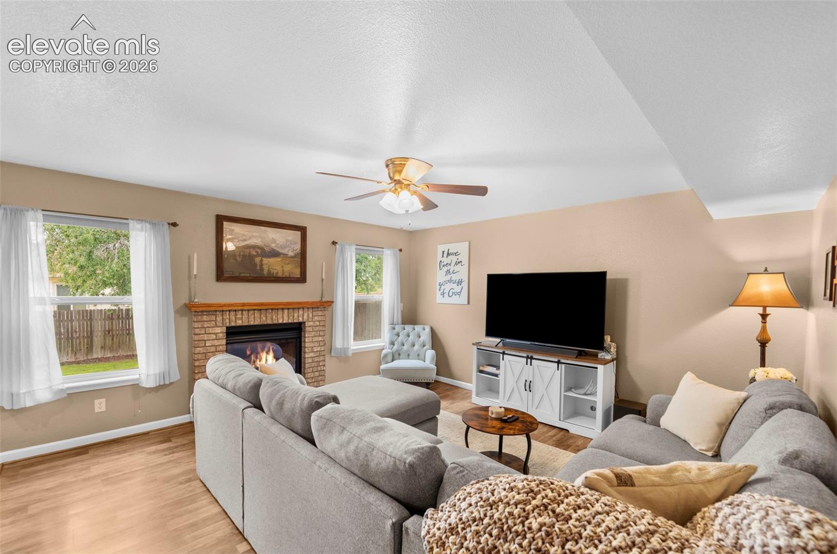 Living area featuring a brick fireplace with a wood mantle, wood-finish flooring, and a ceiling fan