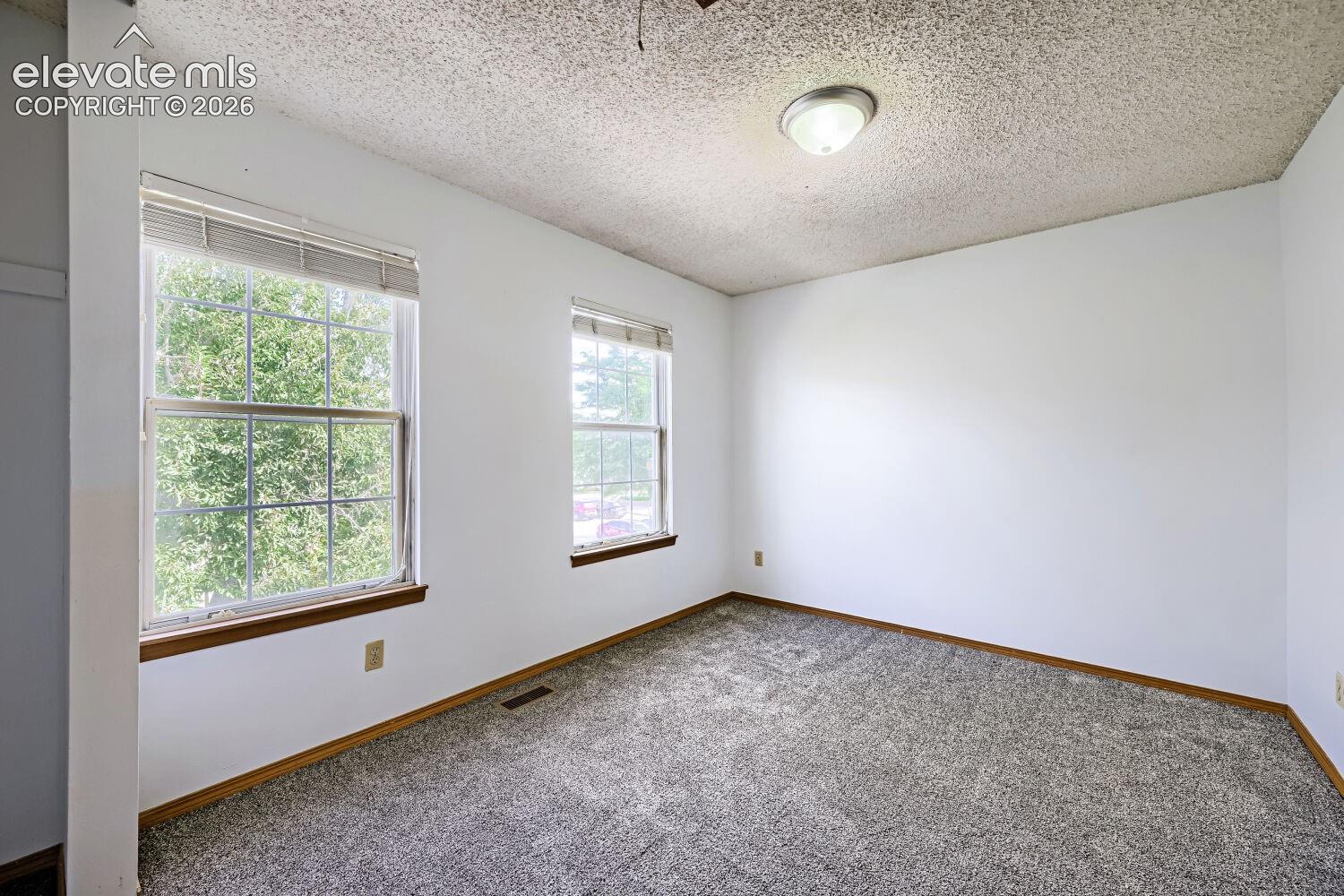 Two large windows, textured ceiling, flush mount lighting, light gray carpeting, and wood trim accents