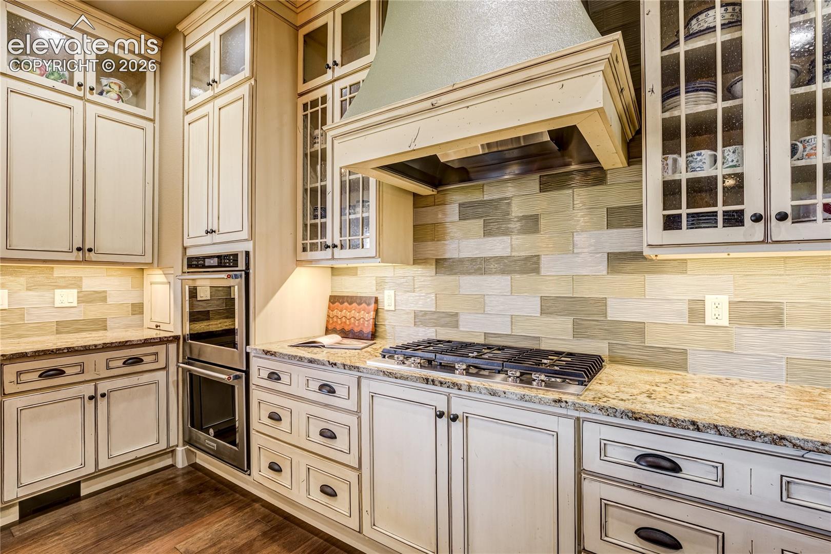 A gas cooktop beneath a decorative vent hood takes center stage, surrounded by custom tilework, detailed cabinetry + integrated double ovens.
