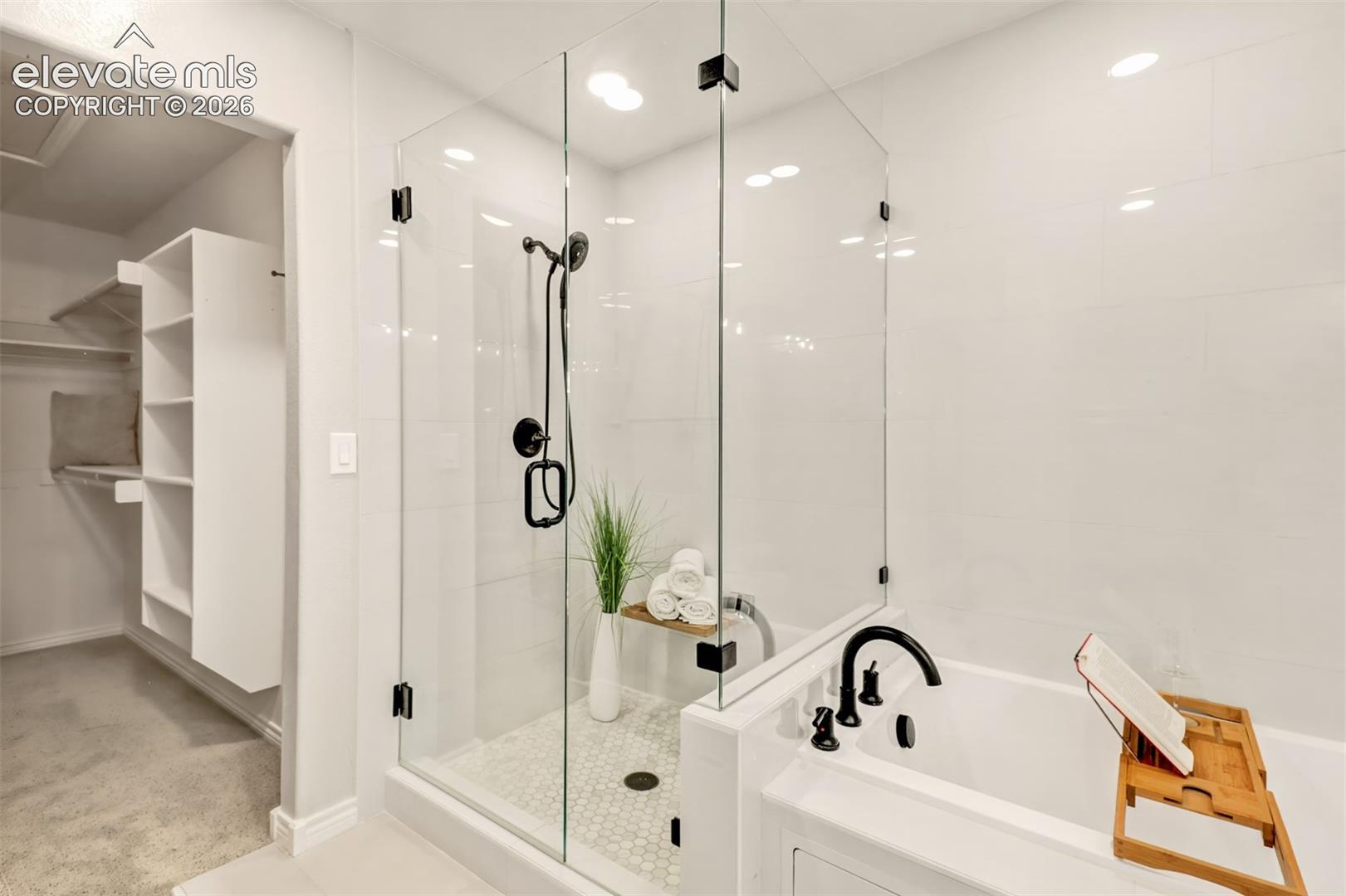 Walk-in glass-enclosed shower featuring a handheld matte black fixture, hexagonal floor tiling, white wall tile, and a soaking tub with a matching matte black faucet