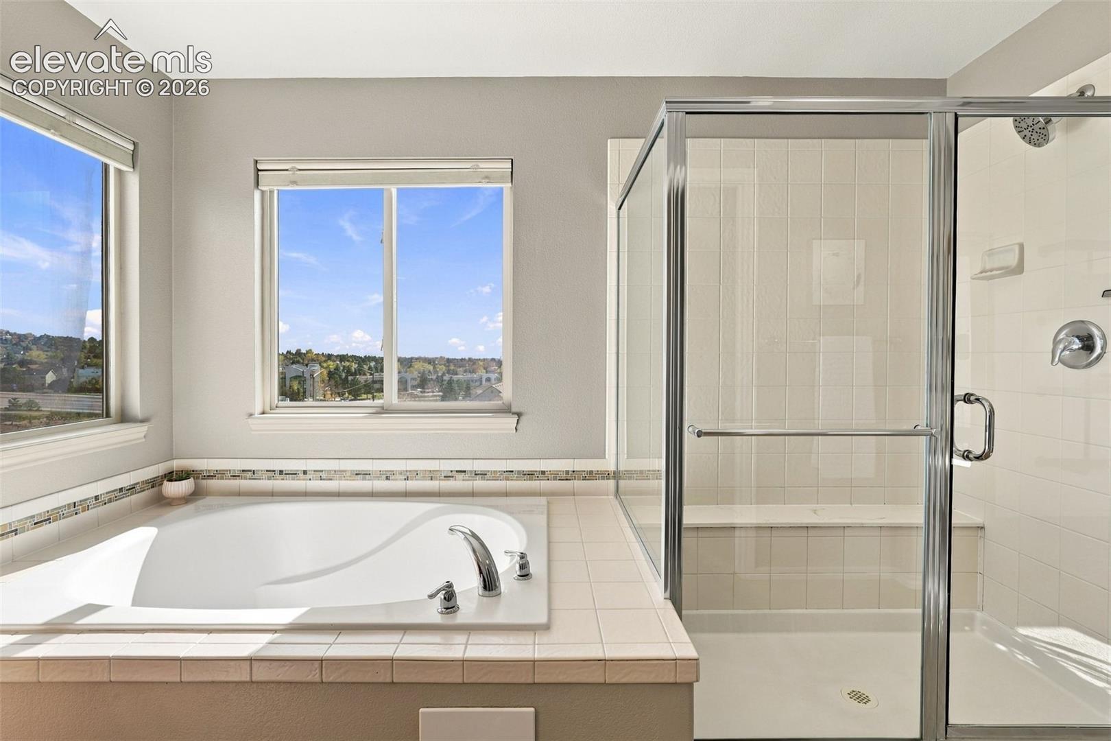 Jetted soaking tub with tile surround, glass-enclosed shower with built-in bench, and dual windows