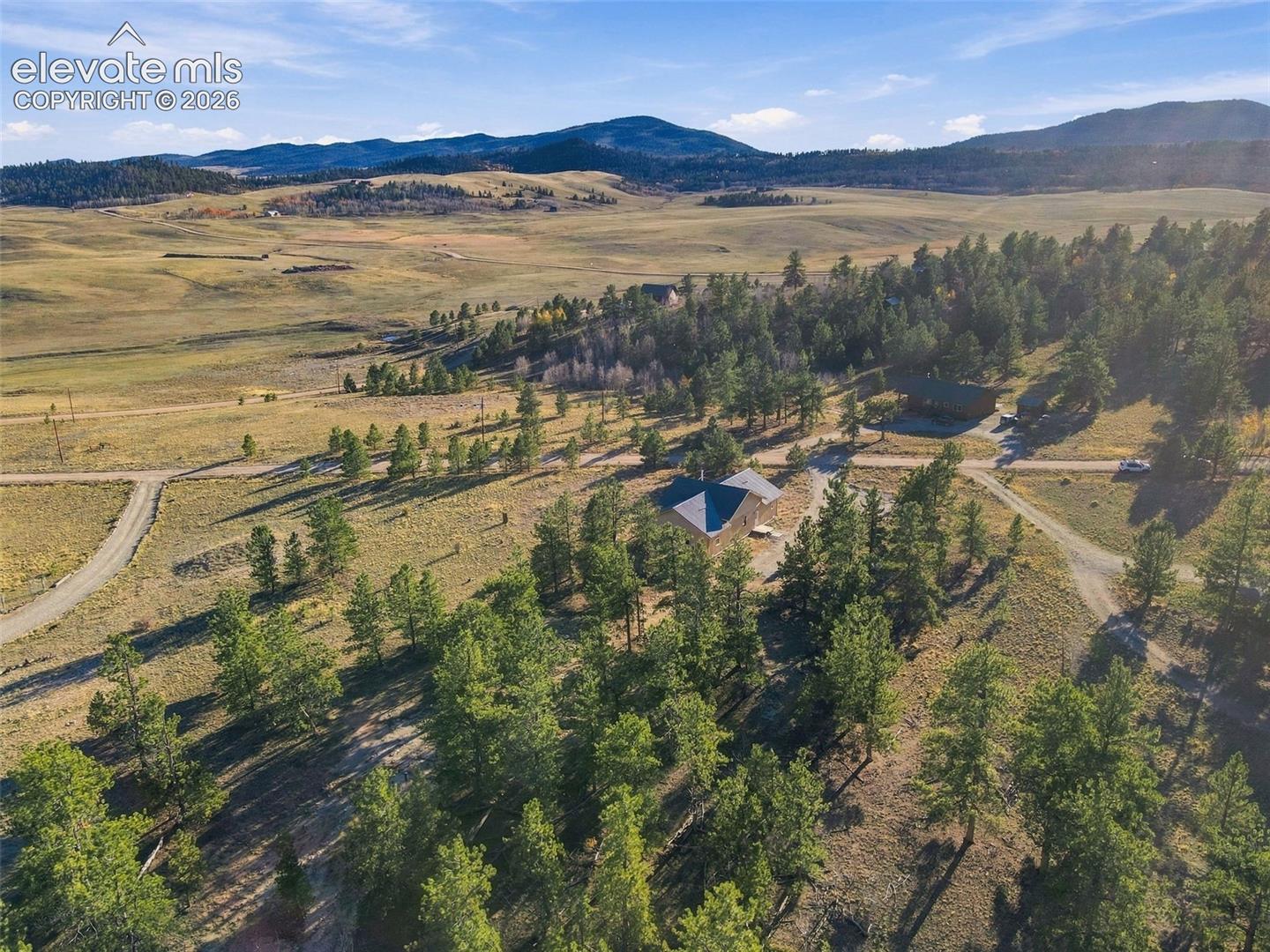 Aerial perspective of a sprawling property featuring mature evergreen trees, expansive open terrain, a multi-gabled roofline, and distant mountain topography