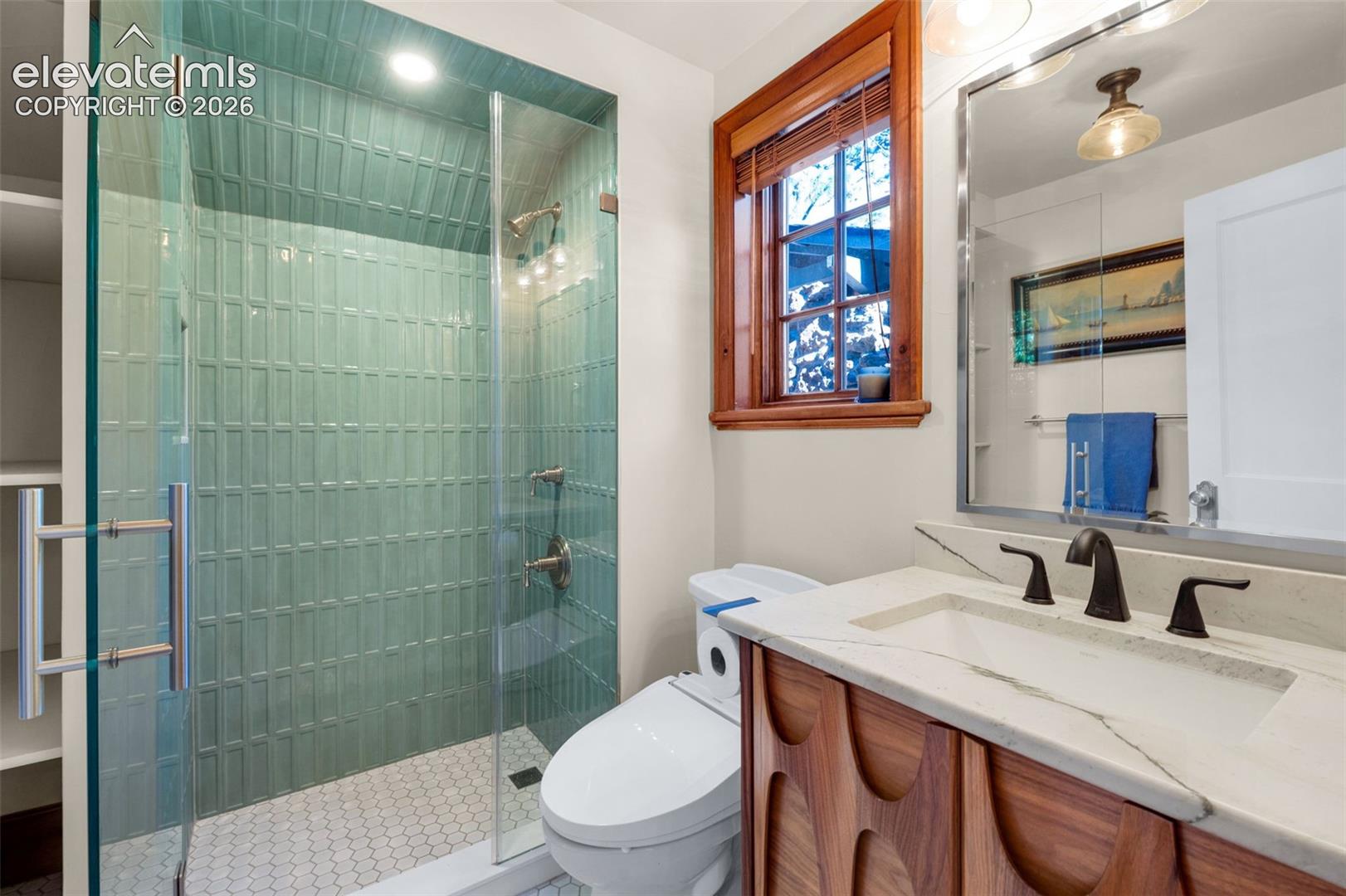 Walk-in shower with vertical teal tile surround and hexagonal floor tile, complemented by a mid-century style wood-finish vanity with a stone countertop, an integrated sink, and an arched-frame mirror
