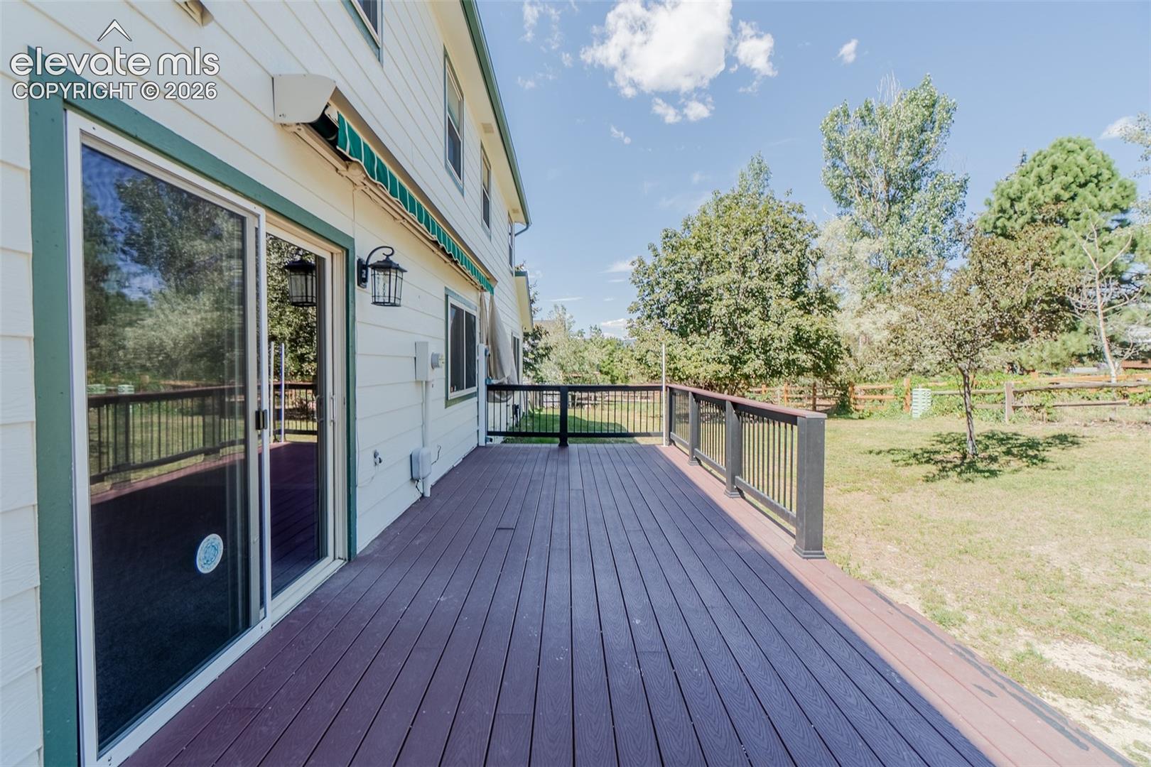 Expansive wood-finish deck with dark railing, featuring a large sliding glass door, a green retractable awning, and exterior wall lantern