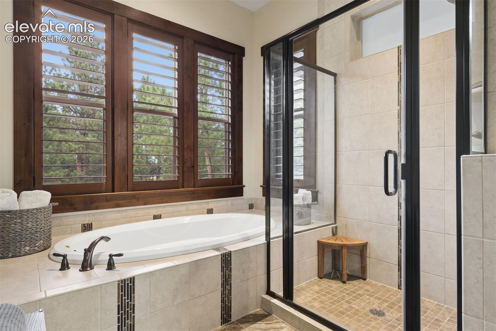 Soaking tub with bronze finish fixtures, integrated tile surround, separate glass-enclosed shower with tiled flooring, and wood-finish window shutters