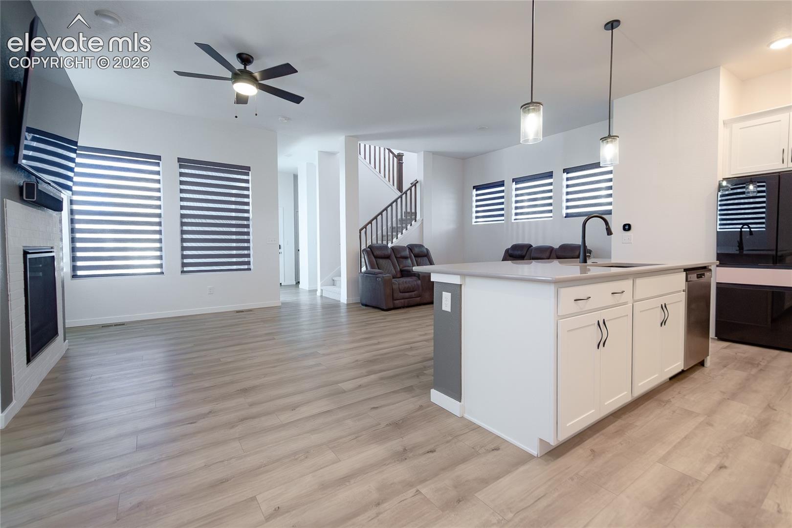 Open-concept interior featuring wood-finish flooring, a kitchen island with neutral-toned countertop and built-in cabinetry, dual pendant lighting, a fireplace with a light-toned surround, and a staircase with a wooden handrail