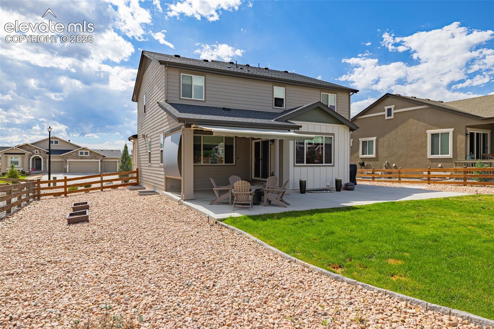 Rear exterior featuring horizontal and board-and-batten siding, a covered concrete patio with a retractable awning, a dedicated gravel landscaping area, and wood-style perimeter fencing
