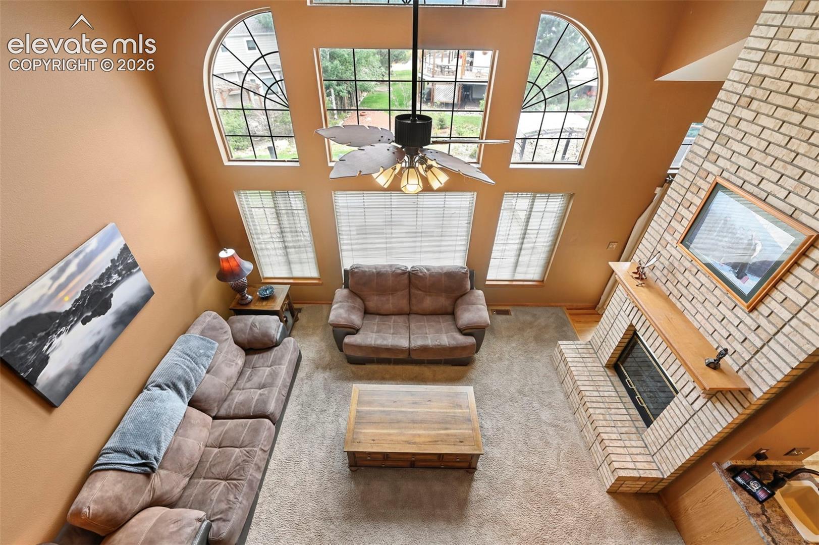 Two-story living area with arched windows, brick-surround fireplace, wood mantel, and ceiling fan with lighting