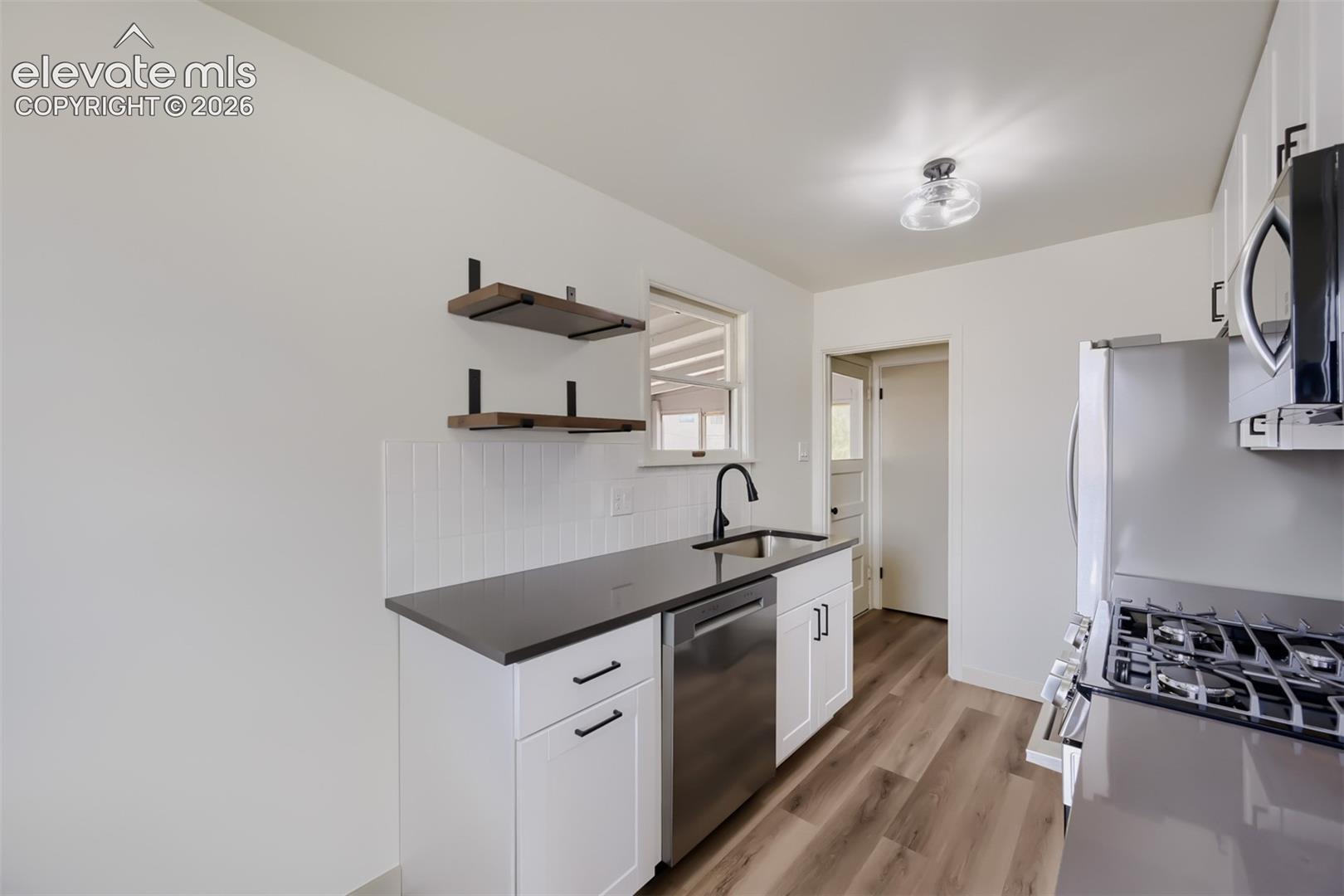Contemporary kitchen featuring white cabinetry, dark countertops, stainless steel appliances, wood-finish flooring, and open shelving